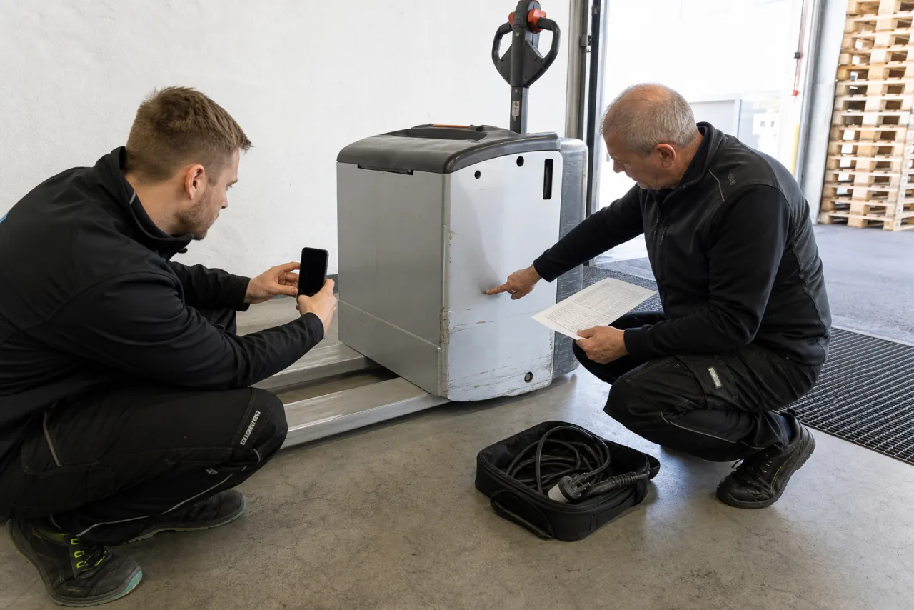 Mitarbeiter fotografiert zusammen mit einem Servicetechniker vorhandene Kratzer und das mitgelieferte Ladekabel eines Miet-Elektrohubwagens.